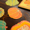Decorative paper pumpkins and leaves on a dark surface with a blurred pumpkin in the background. Squash Autumn Handprinted Garland | East End Press | Shop Fodder