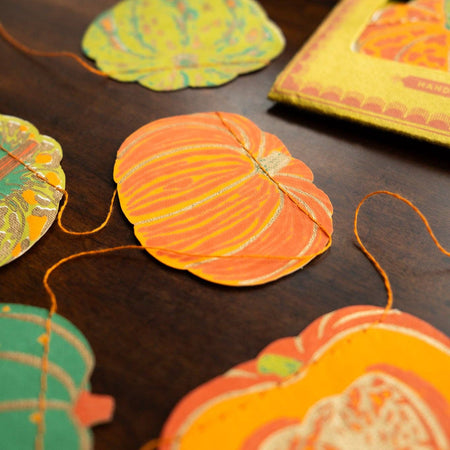 Decorative paper pumpkins and leaves on a dark surface with a blurred pumpkin in the background. Squash Autumn Handprinted Garland | East End Press | Shop Fodder