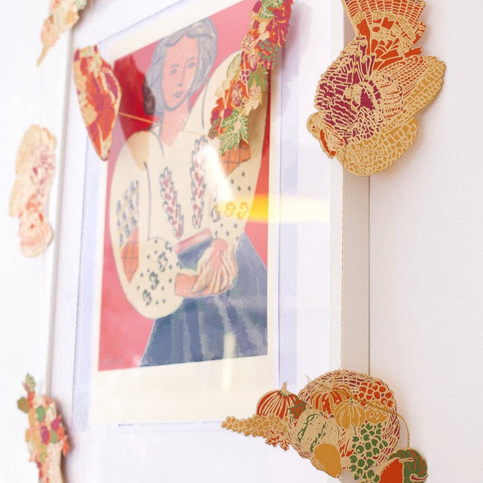 Decorative paper flowers hanging on a white wall with a colorful poster in the background. Thanksgiving Handprinted Garland | East End Press | Shop Fodder