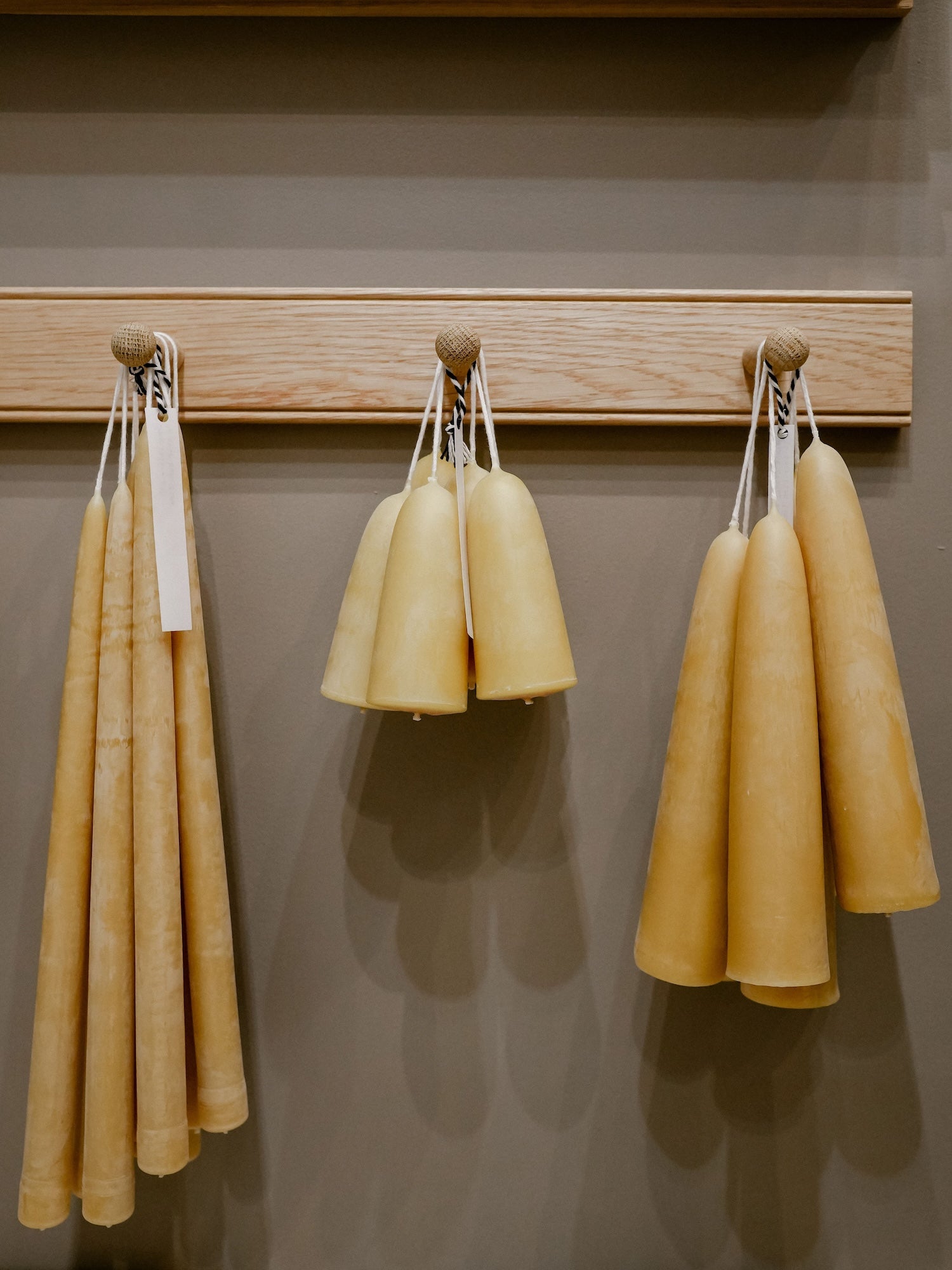 Three natural beeswax candles in tapered shapes hang from wooden pegs on a light wood rack against a warm taupe wall.