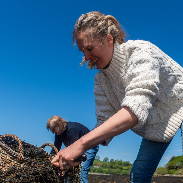 Woman in a white sweater handling seaweed with another person in the background under a clear blue sky. | Seaweed Foot Scrub | WASI Seaweed | Shop Fodder