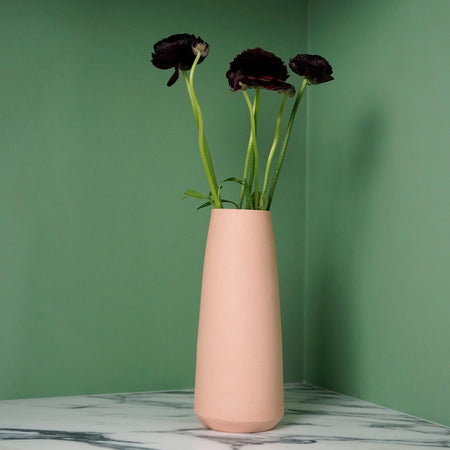 a pink tall tapered vase on a marble table against a bright green background with five deep red flowers. 