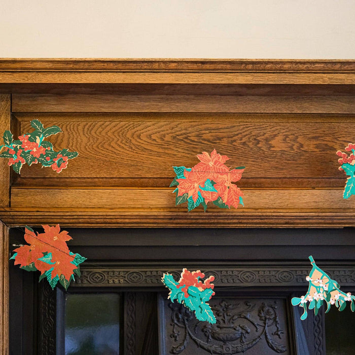 Decorative paper flowers on a wooden mantelpiece with a Christmas tree in the background. East End Press | Shop Fodder