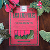 Box of East End Press ornaments with decorative paper holly leaves and berries on a wooden surface. East End Press | Shop Fodder