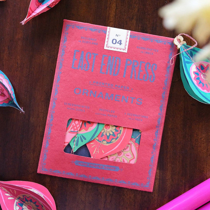 Red packaging of East End Press ornaments on a wooden surface with decorative items around. East End Press | Shop Fodder