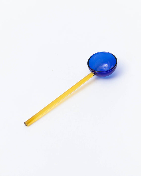 A translucent glass ladle with a vibrant blue bowl and a long, slender yellow handle, photographed on a seamless white background.