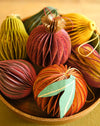 A group of decorative handmade paper ornaments shaped like pumpkins or gourds, arranged in a wooden bowl on a warm neutral surface. The ornaments feature layered paper pleats in shades of rust red, orange, yellow, green, and teal with thin string details.