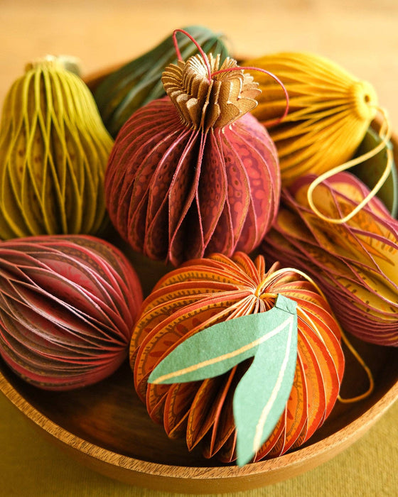 A group of decorative handmade paper ornaments shaped like pumpkins or gourds, arranged in a wooden bowl on a warm neutral surface. The ornaments feature layered paper pleats in shades of rust red, orange, yellow, green, and teal with thin string details.